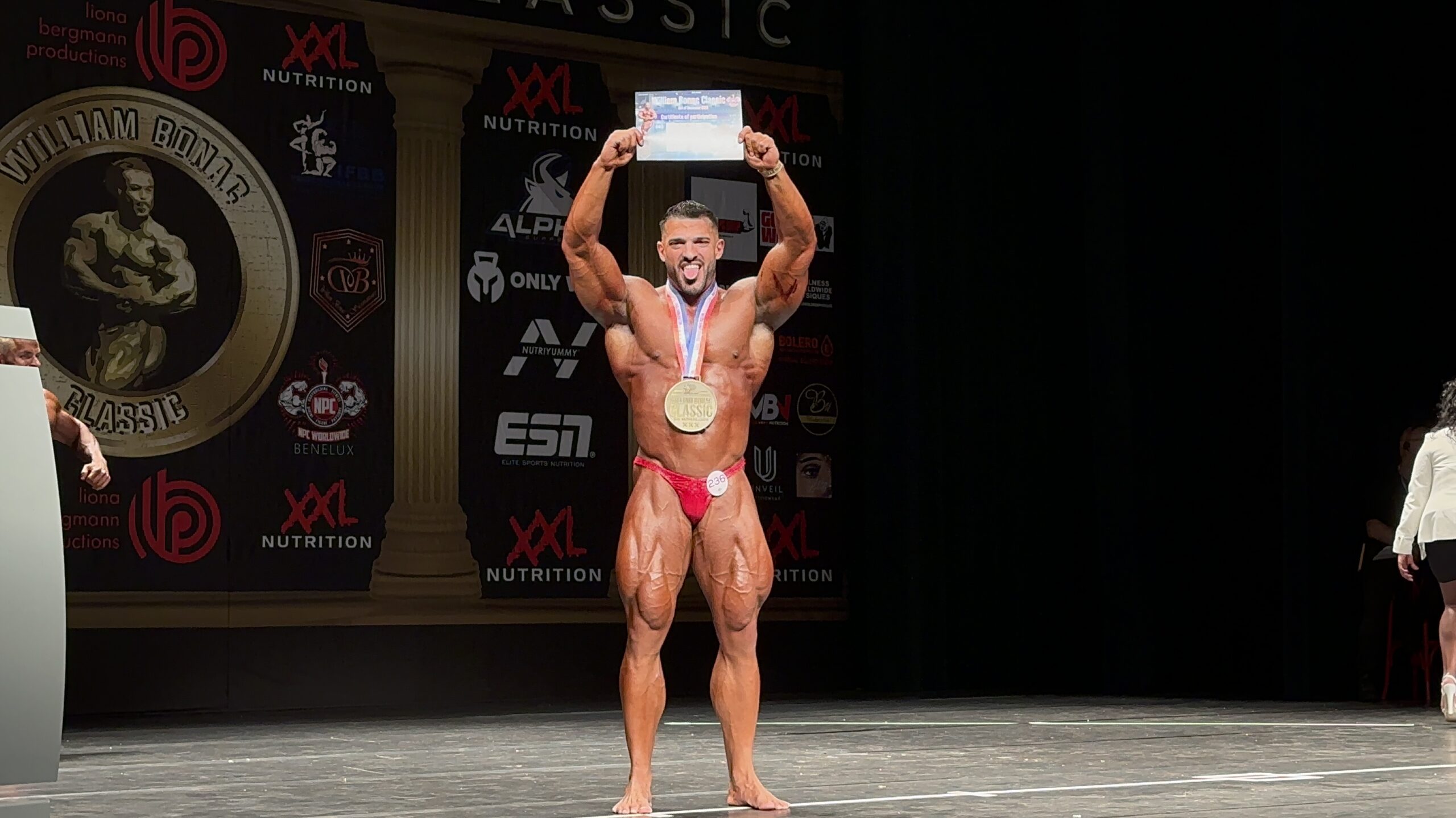 Lars Gorilla, competitive bodybuilder and LANCEGYM online coaching collaborator, celebrating on stage with a medal and certificate, showcasing his muscular physique.